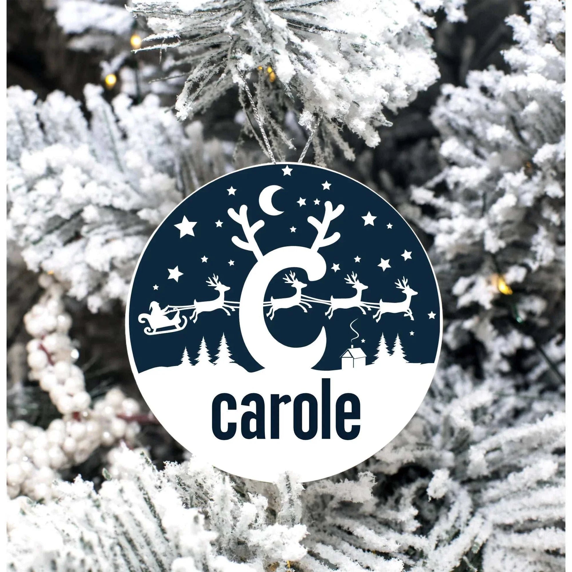 Monogrammed round porcelain Christmas ornament with reindeer scene and personalized name hanging on snowy tree branch