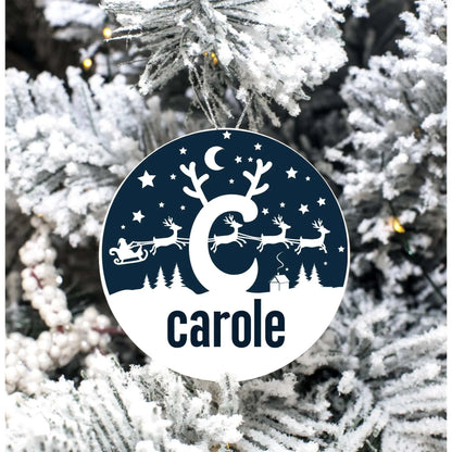 Monogrammed round porcelain Christmas ornament with reindeer scene and personalized name hanging on snowy tree branch