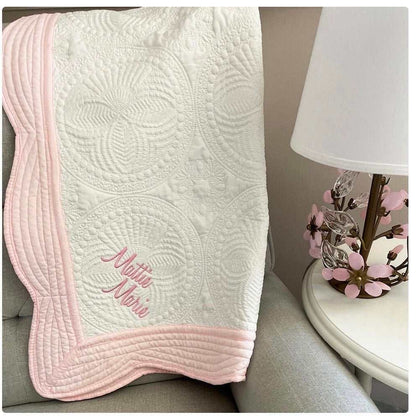 Personalized embroidered baby quilt with pink scalloped edges on a chair near a floral lamp.