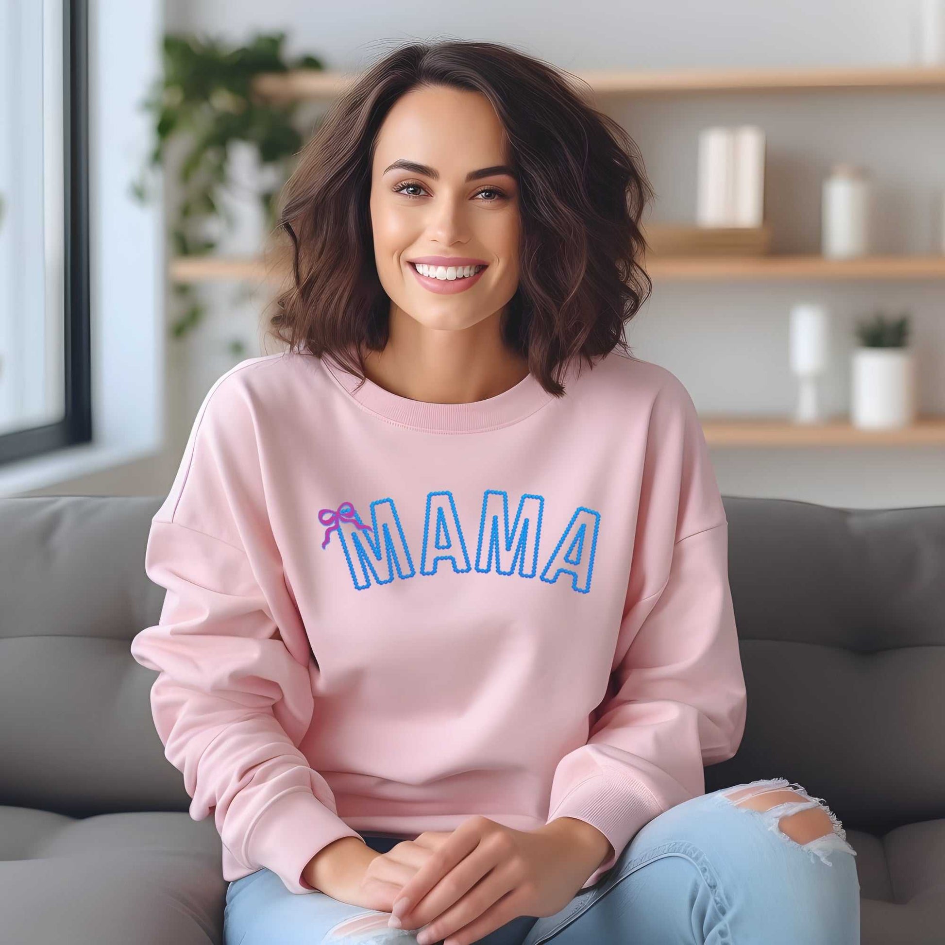 Custom embroidered "MAMA" sweatshirt in pink with blue thread and bow detail, worn by a woman sitting on a sofa.