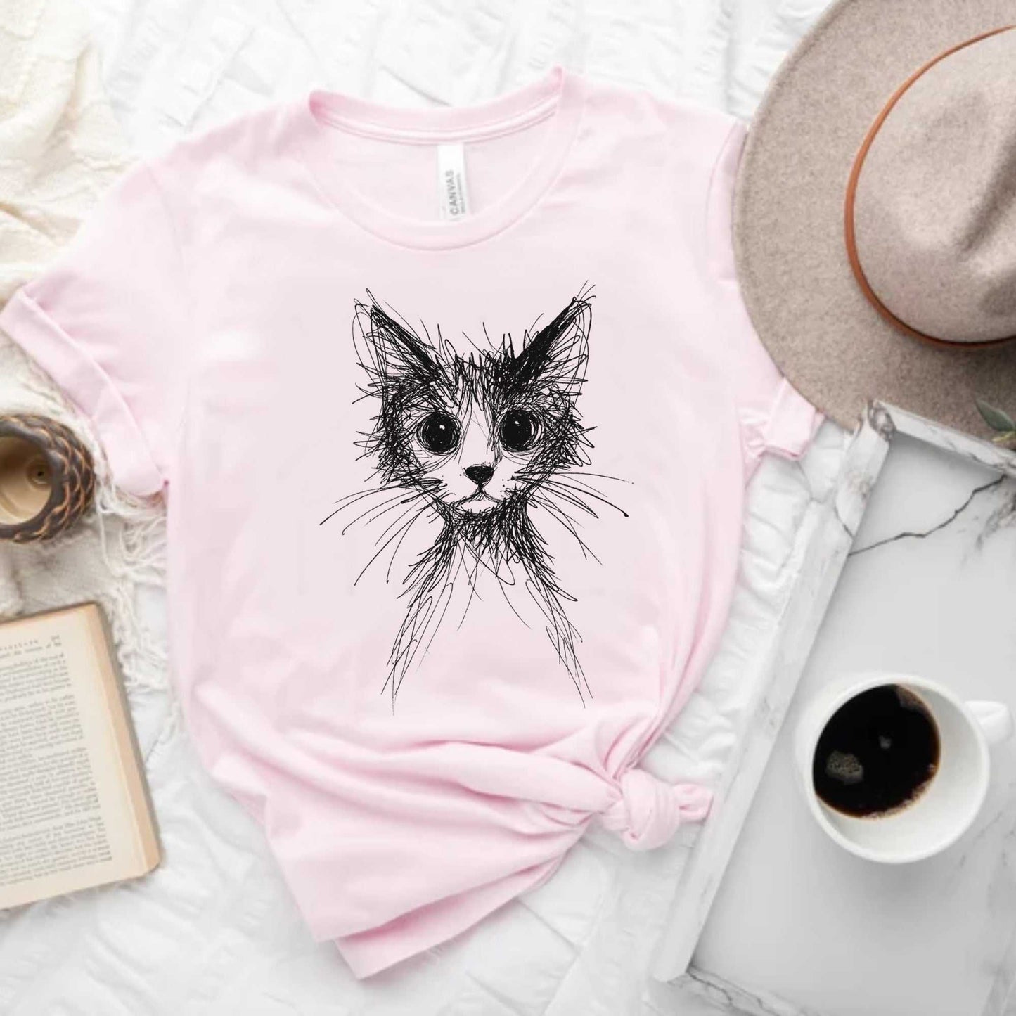 Pink t-shirt with a black cat illustration on a white surface with a hat, book, and coffee cup.