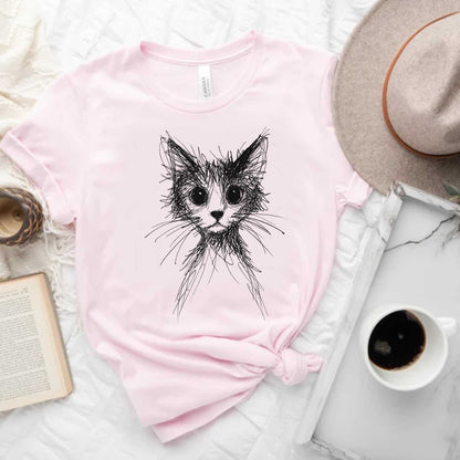 Pink t-shirt with a black cat illustration on a white surface with a hat, book, and coffee cup.