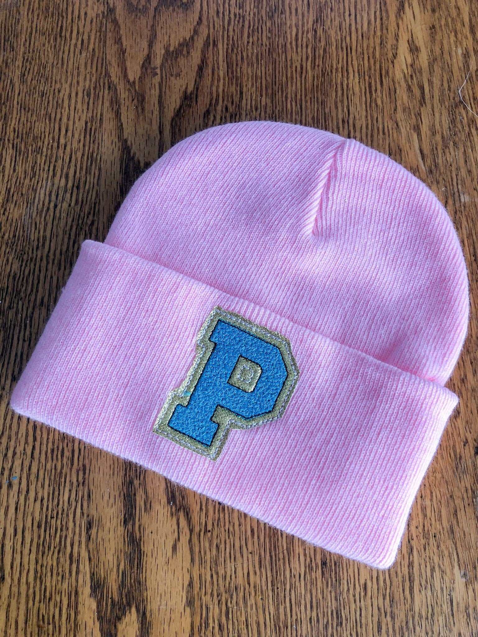 Custom pink embroidered initial beanie with blue 'P' on wooden surface.