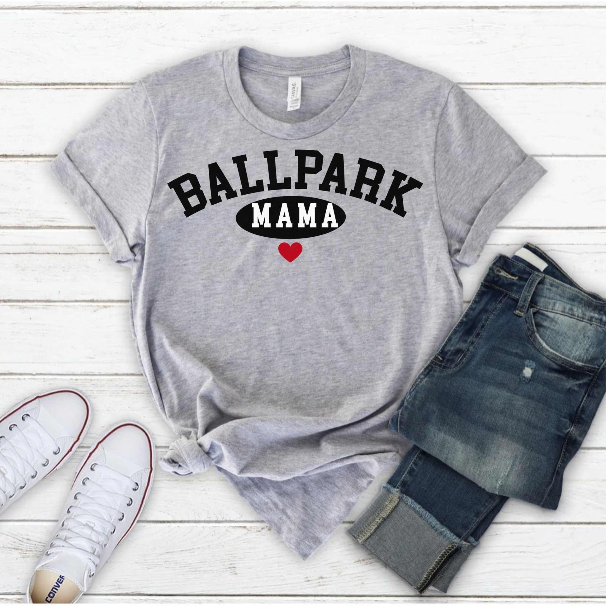 Ballpark Mama Baseball T-Shirt with bold black text and red heart design, displayed with casual sneakers and jeans against a wooden