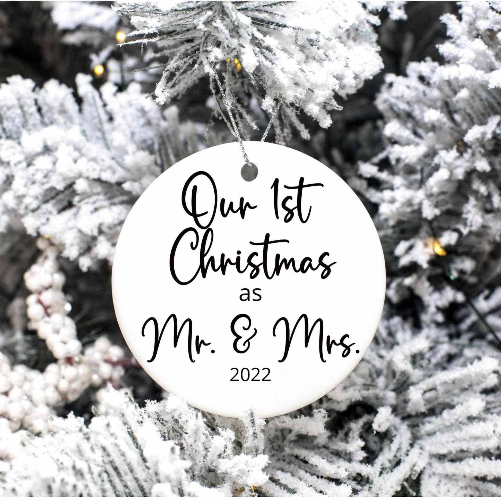 Personalized round porcelain Christmas ornament reading "Our 1st Christmas as Mr. & Mrs. 2022" hanging on snowy tree branch with gold string.