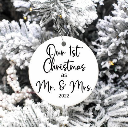 Personalized round porcelain Christmas ornament reading "Our 1st Christmas as Mr. & Mrs. 2022" hanging on snowy tree branch with gold string.