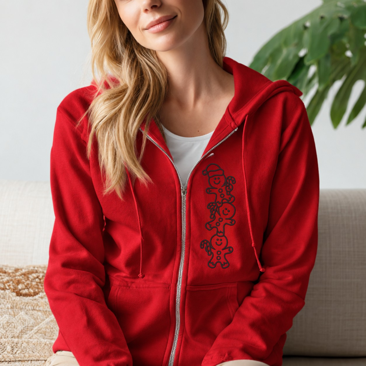 A woman wearing a bright red zip-up hoodie with a black stacked gingerbread cookie graphic on the left chest, sitting on a beige couch.