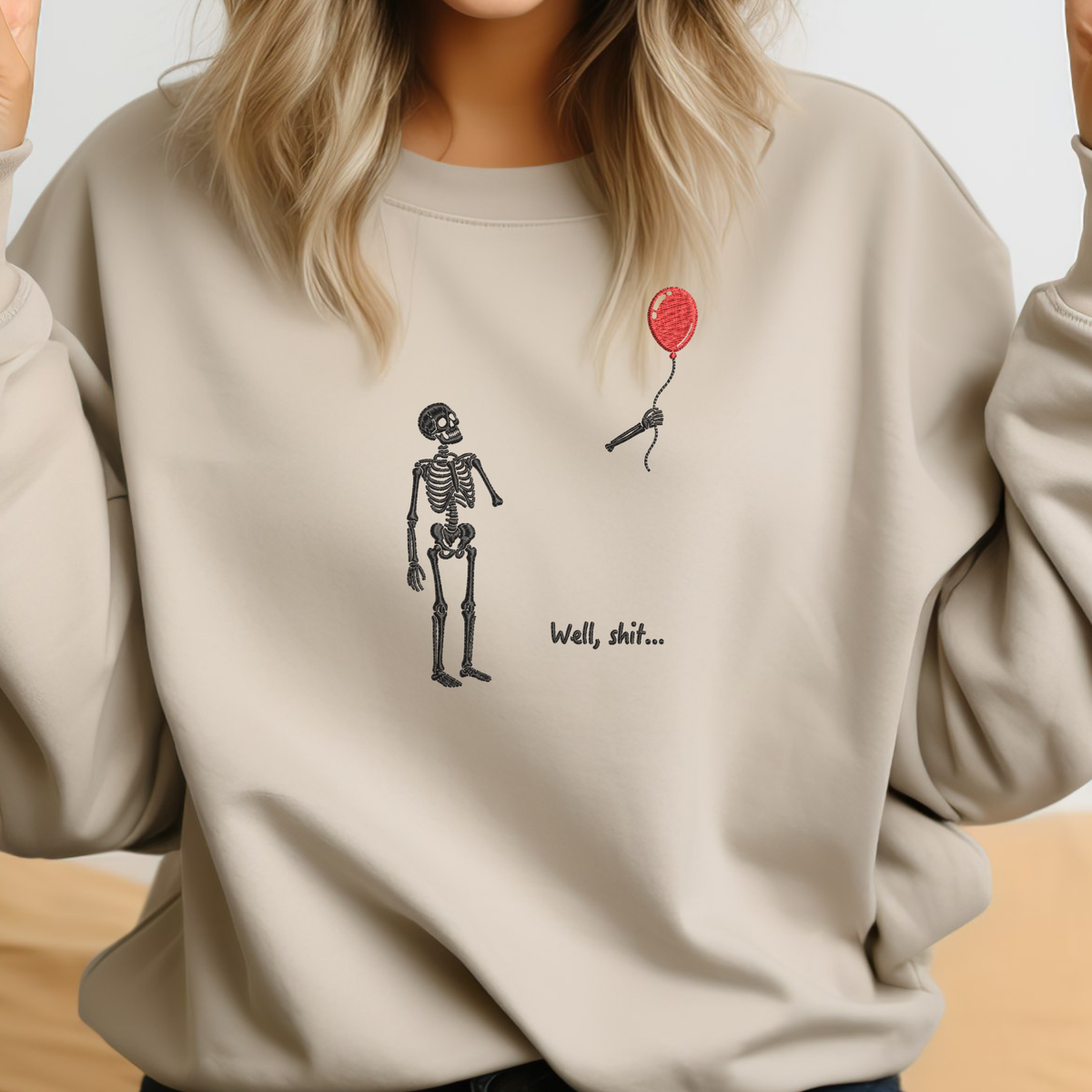 A woman wearing a beige crewneck sweatshirt with a graphic of a black skeleton standing next to a red balloon and the text 'Well, shit.