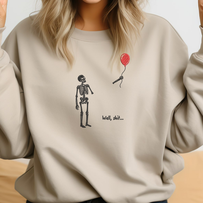 A woman wearing a beige crewneck sweatshirt with a graphic of a black skeleton standing next to a red balloon and the text 'Well, shit.