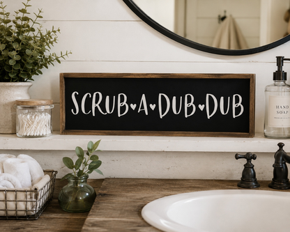 Bathroom vanity with 'Scrub A Dub Dub' sign, plants, and soap dispenser.