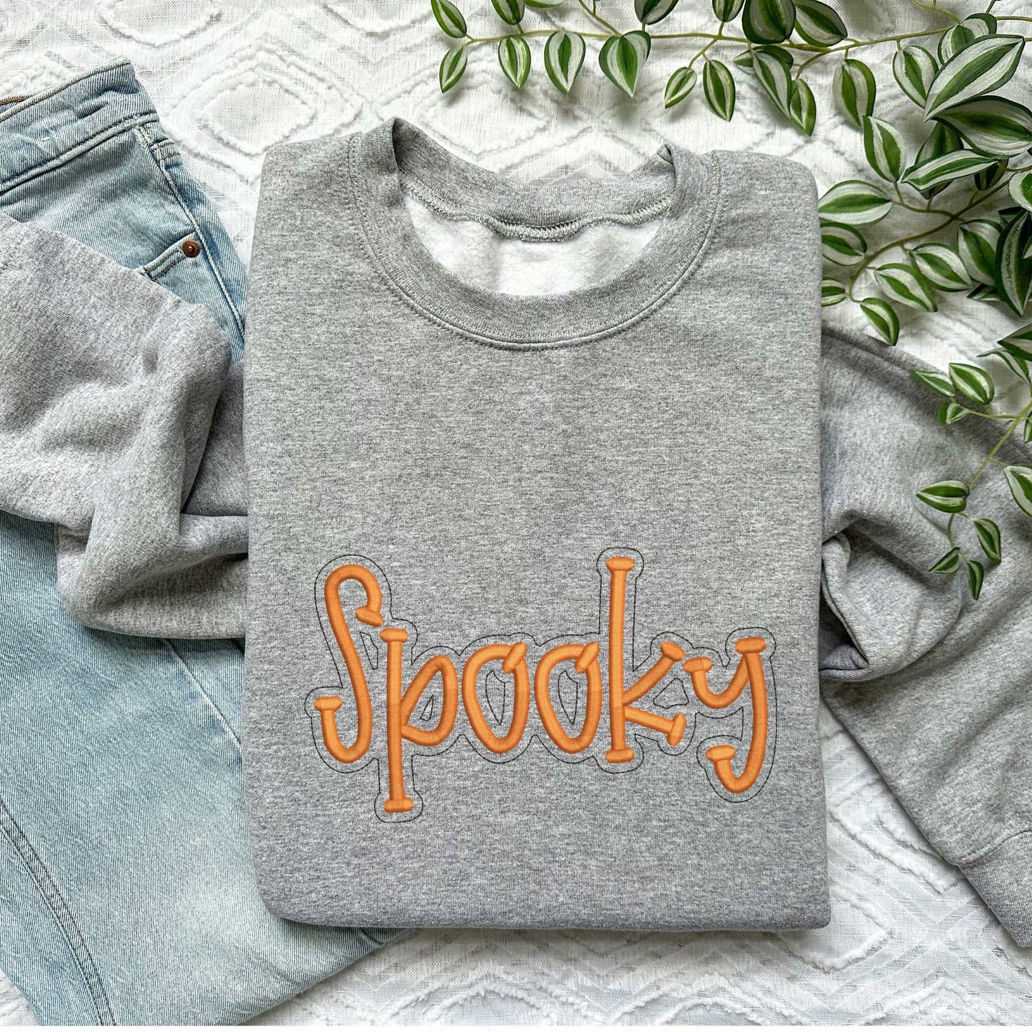 Gray crewneck sweater with orange embroidered "Spooky" text, styled for fall and Halloween, laid on a light background next to blue jeans and green plant leaves.