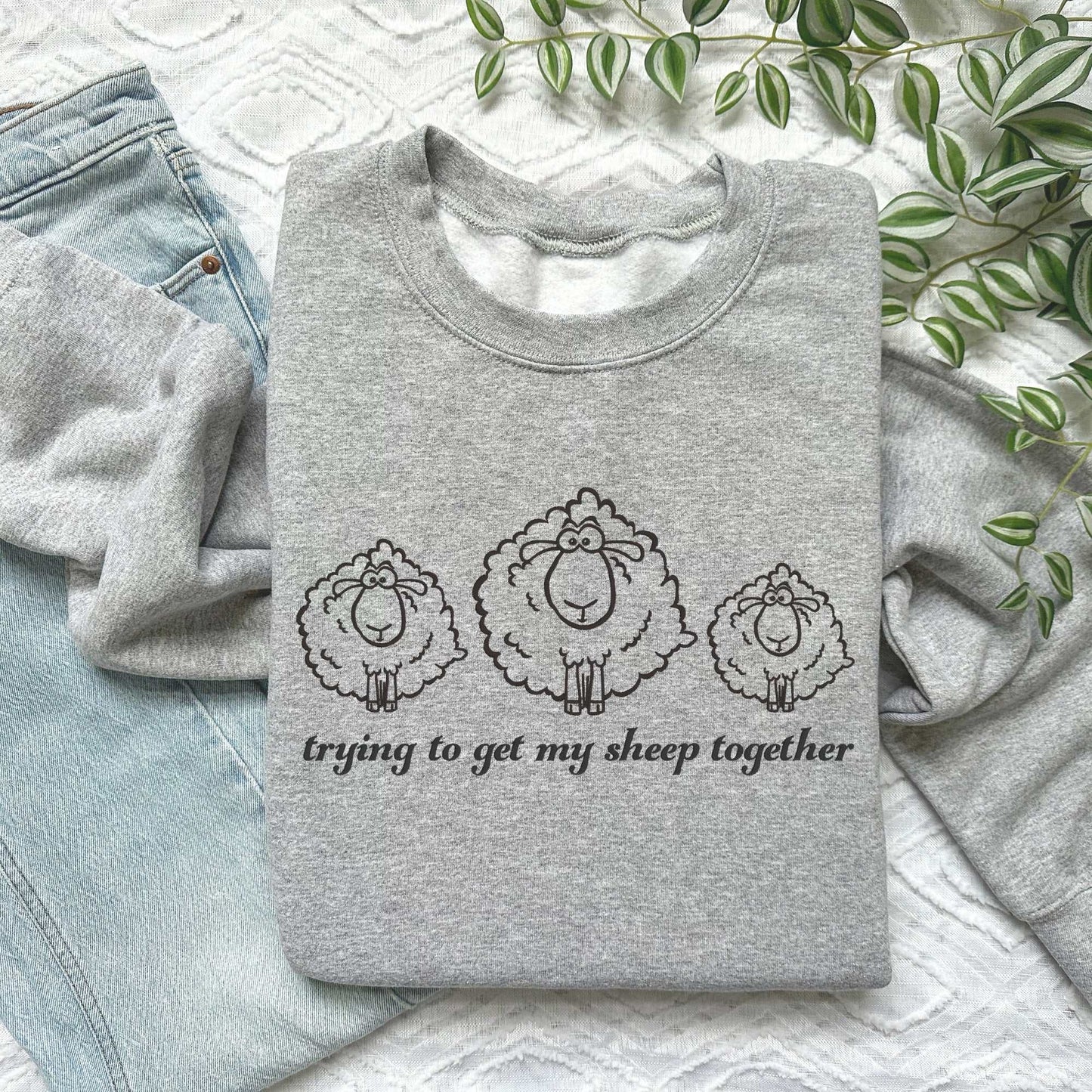 Gray embroidered sweatshirt with three fluffy sheep and "Trying to Get My Sheep Together" text, laid on jeans.