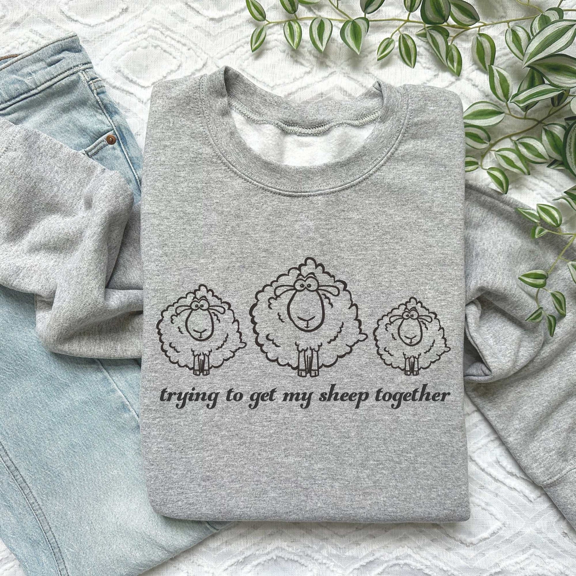 Gray embroidered sweatshirt with three fluffy sheep and "Trying to Get My Sheep Together" text, laid on jeans.