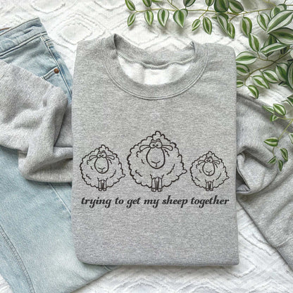 Gray embroidered sweatshirt with three fluffy sheep and "Trying to Get My Sheep Together" text, laid on jeans.
