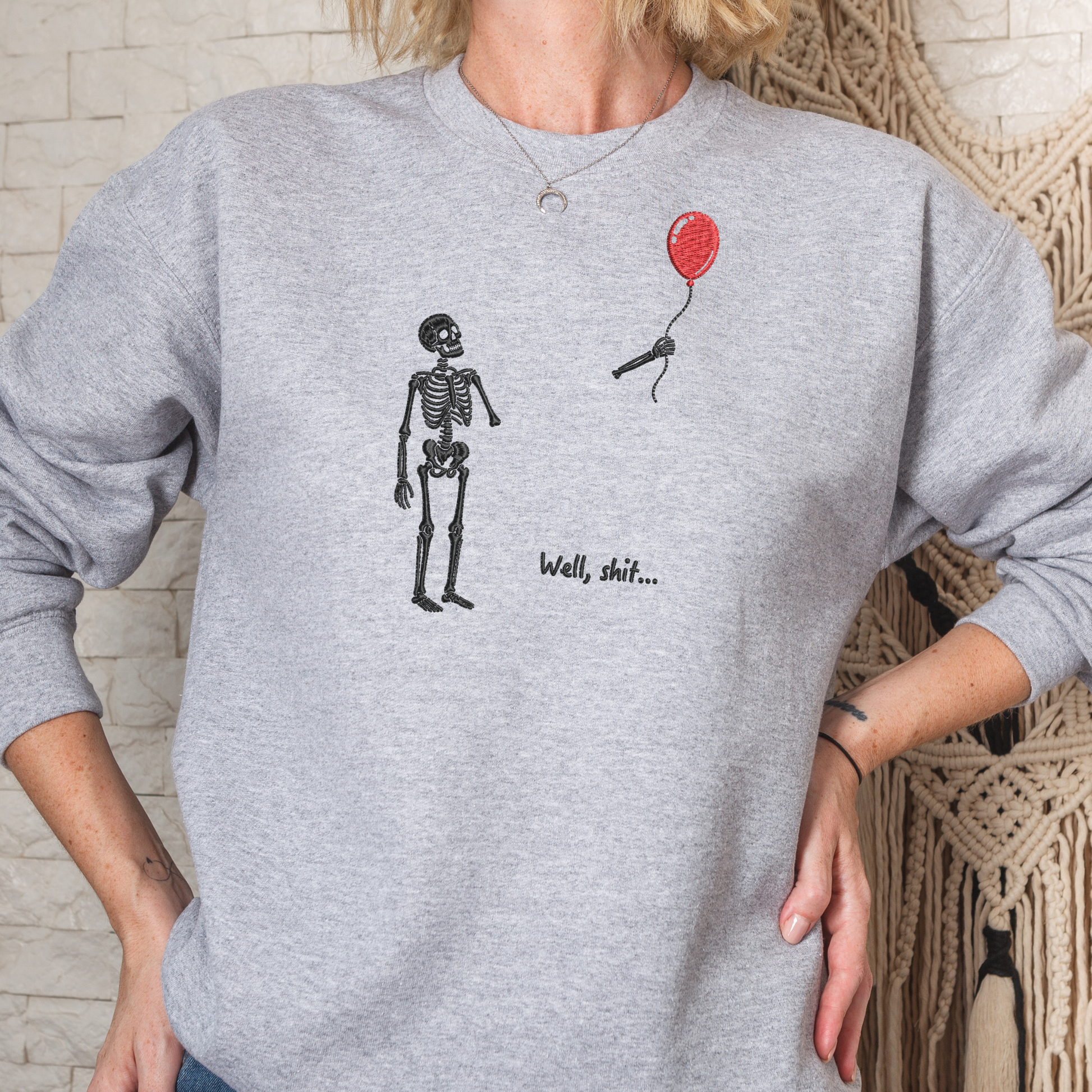 A person wearing a heather gray crewneck sweatshirt with a graphic of a black skeleton standing next to a small red balloon and the text 'Well, shit.