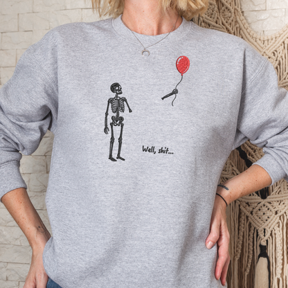 A person wearing a heather gray crewneck sweatshirt with a graphic of a black skeleton standing next to a small red balloon and the text 'Well, shit.