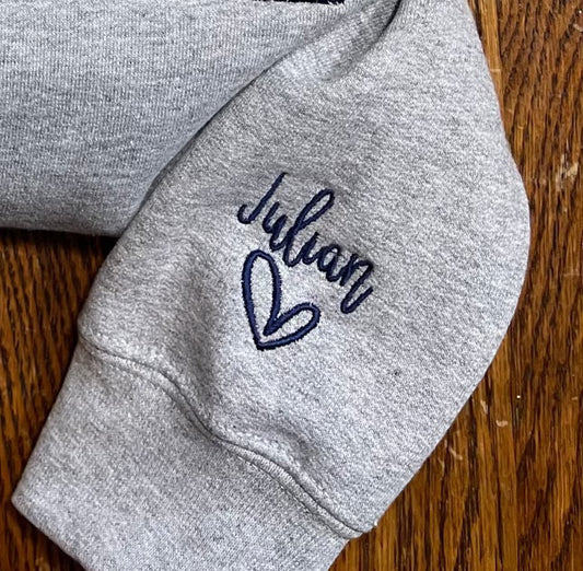 Gray sweater sleeve with navy blue embroidered name "Julian" and heart design
