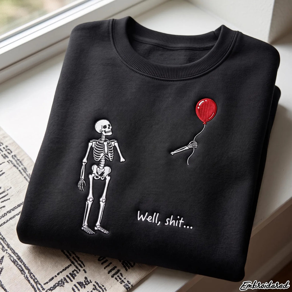 A folded black crewneck sweatshirt with a white skeleton graphic, a red balloon, and the text 'Well, shit.