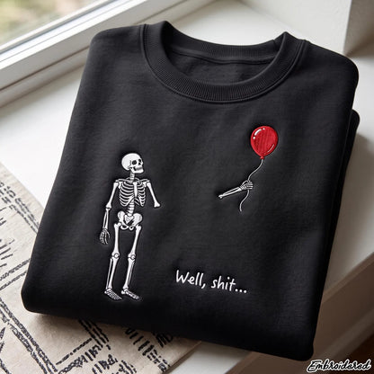 A folded black crewneck sweatshirt with a white skeleton graphic, a red balloon, and the text 'Well, shit.