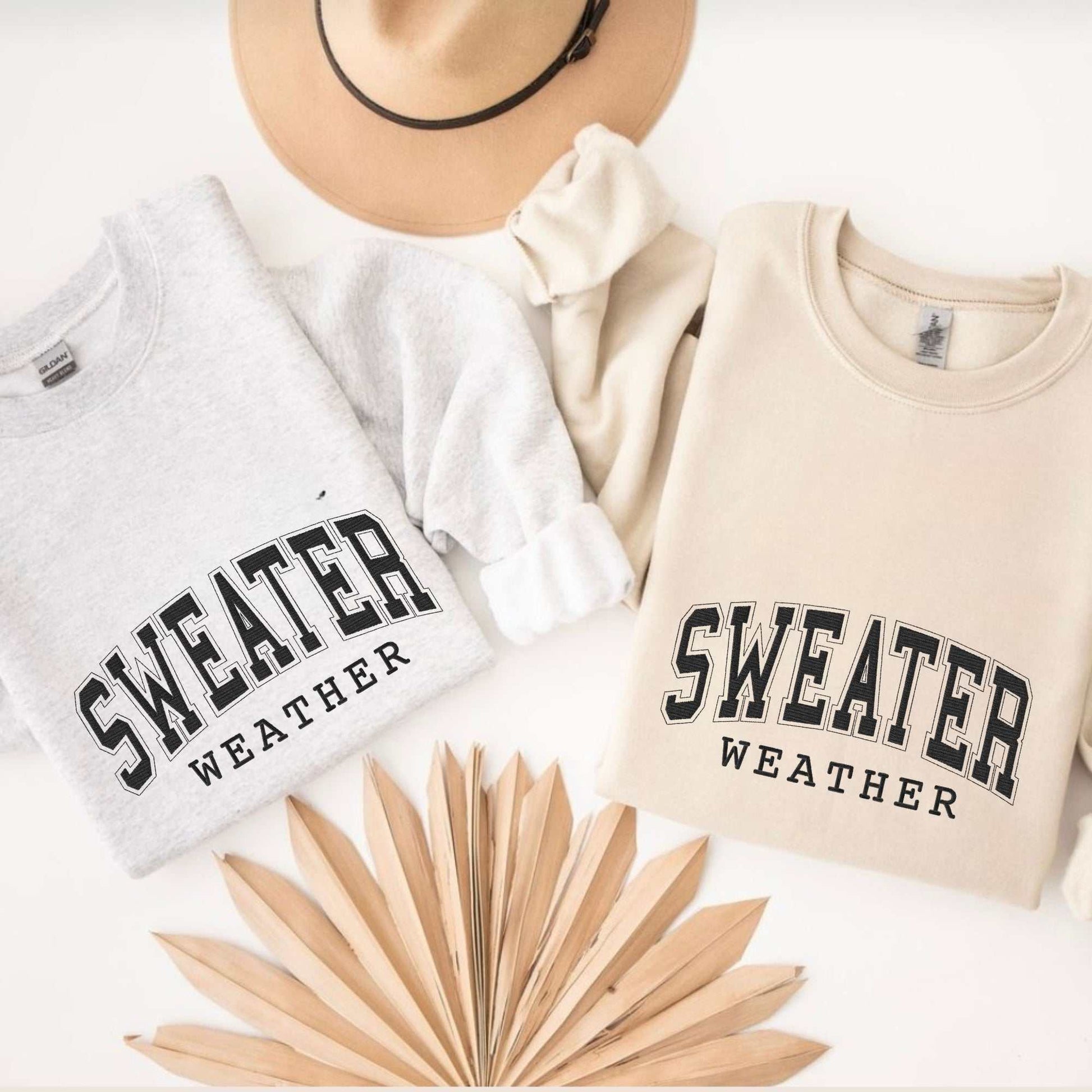 Two cozy embroidered crewneck sweatshirts in light gray and beige with "Sweater Weather" text, styled with a tan hat and dried palm leaves for fall fashion.