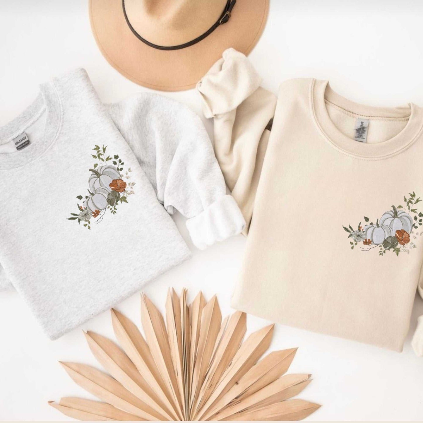 Two folded white floral pumpkin embroidered sweaters in light gray and beige with a tan hat and dried palm leaves on a white background