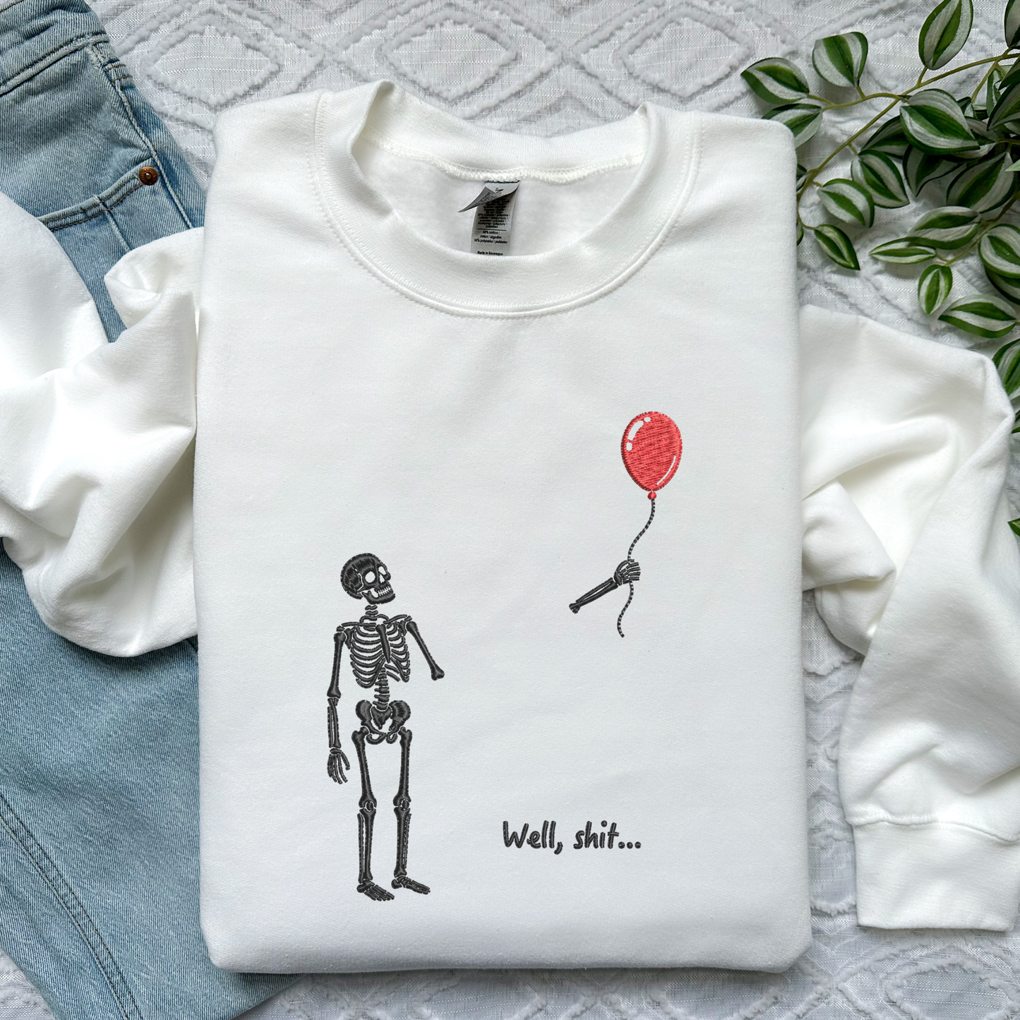 A folded white long-sleeve sweatshirt with a graphic of a skeleton looking at a red balloon and the text 'Well, shit.