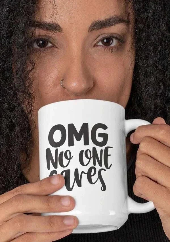 White ceramic coffee mug with bold black "OMG No One Cares" typography, held by person, sarcastic office drinkware.