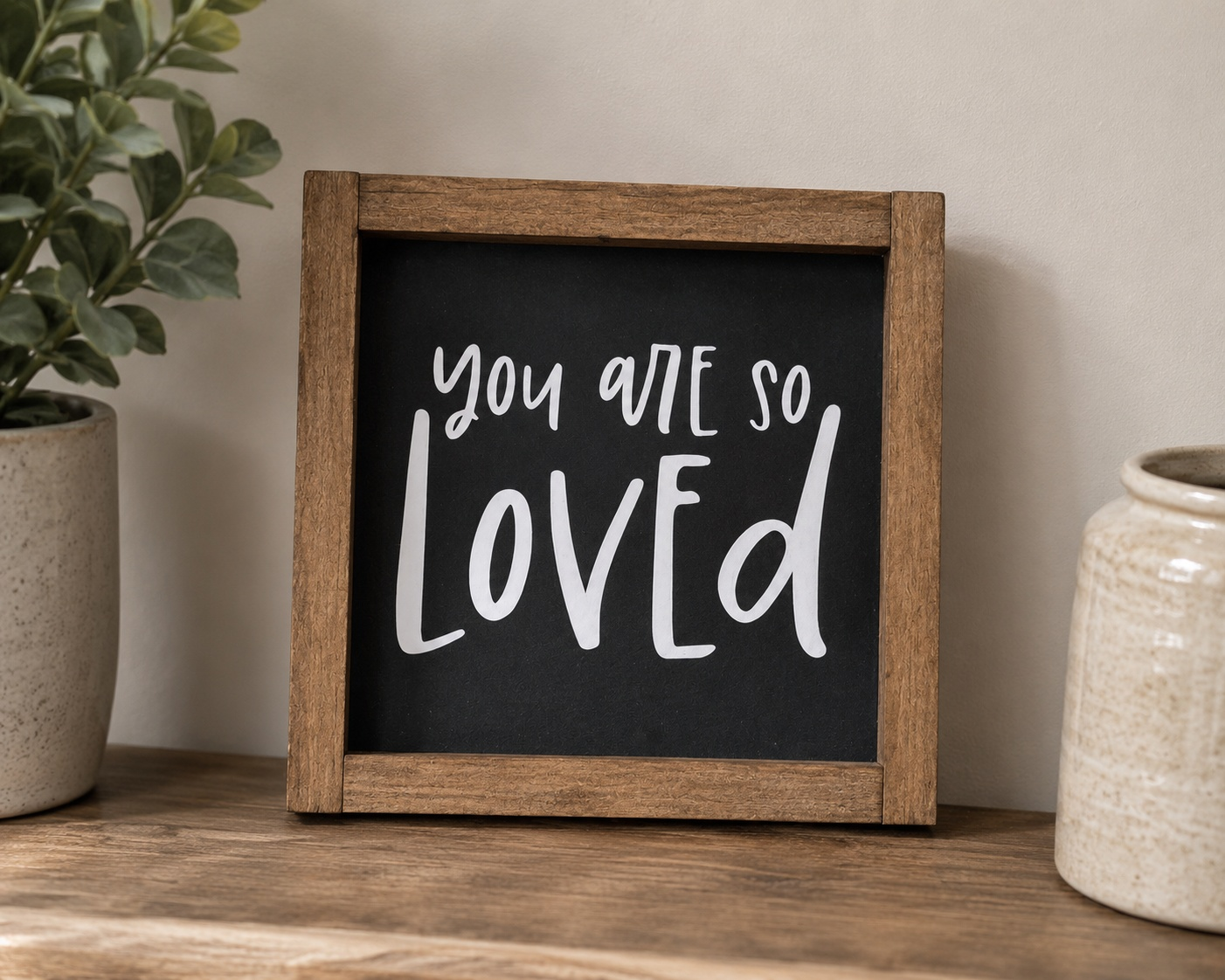 Framed sign with 'You are so loved' text on a wooden surface with plants and a vase.