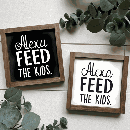 Handcrafted "Alexa, Feed The Kids" framed wood sign with black and white lettering, surrounded by greenery.