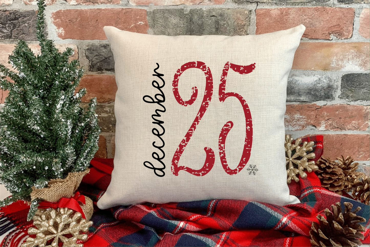 Christmas pillow cover with "December 25" grunge design on natural cotton canvas, placed on red plaid blanket, next to a small Christmas tree and pinecones.