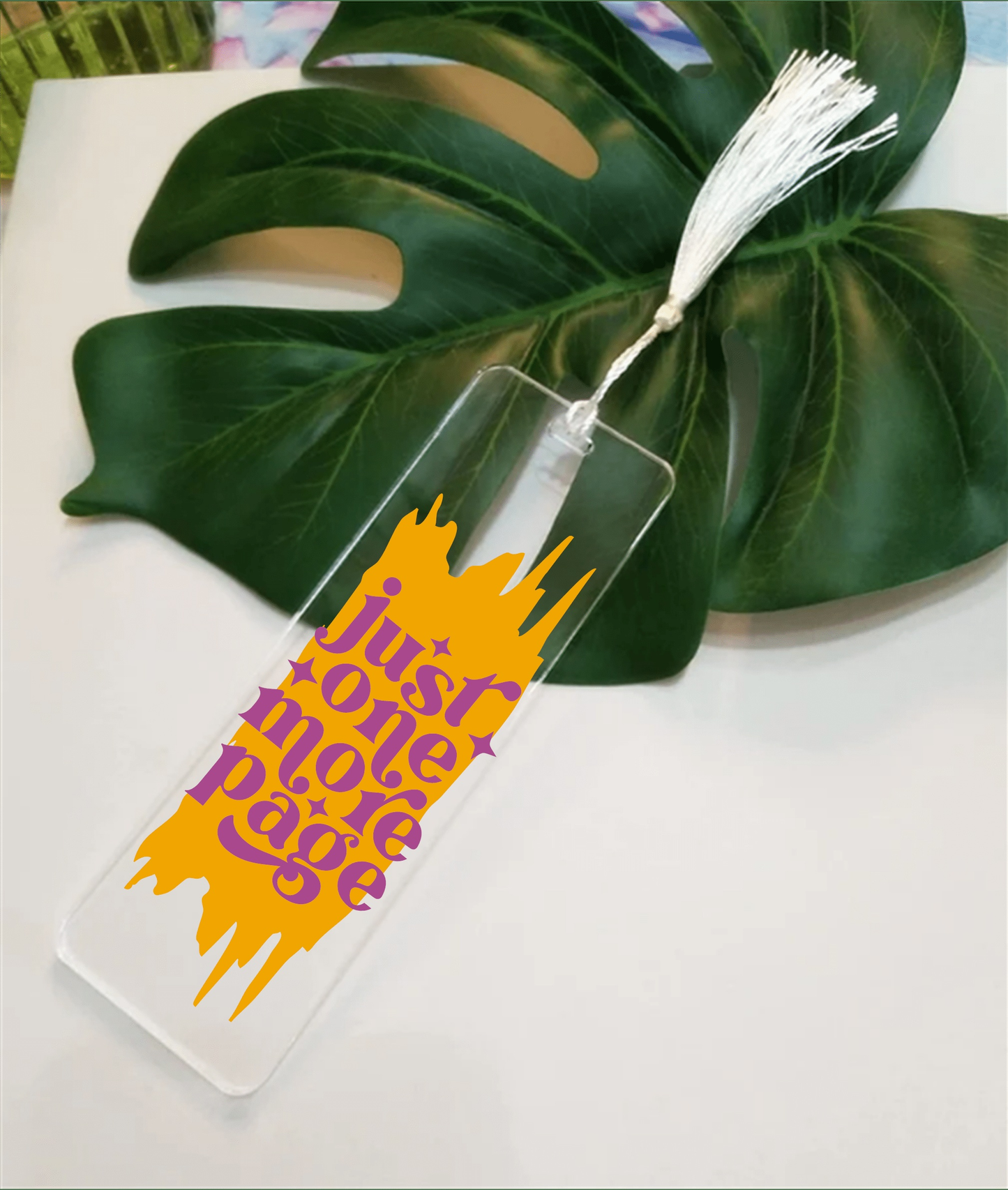 Acrylic bookmark "Just One More Page" with tassel displayed on a leaf background.