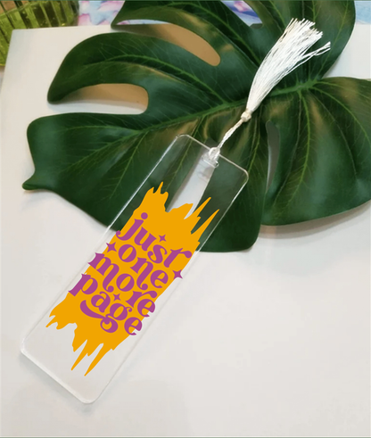 Acrylic bookmark "Just One More Page" with tassel displayed on a leaf background.