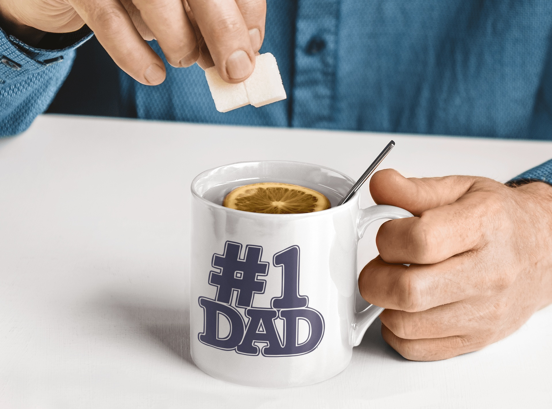 White mug with blue #1 Dad sticker being held by a man adding sugar to a hot drink with lemon slice.