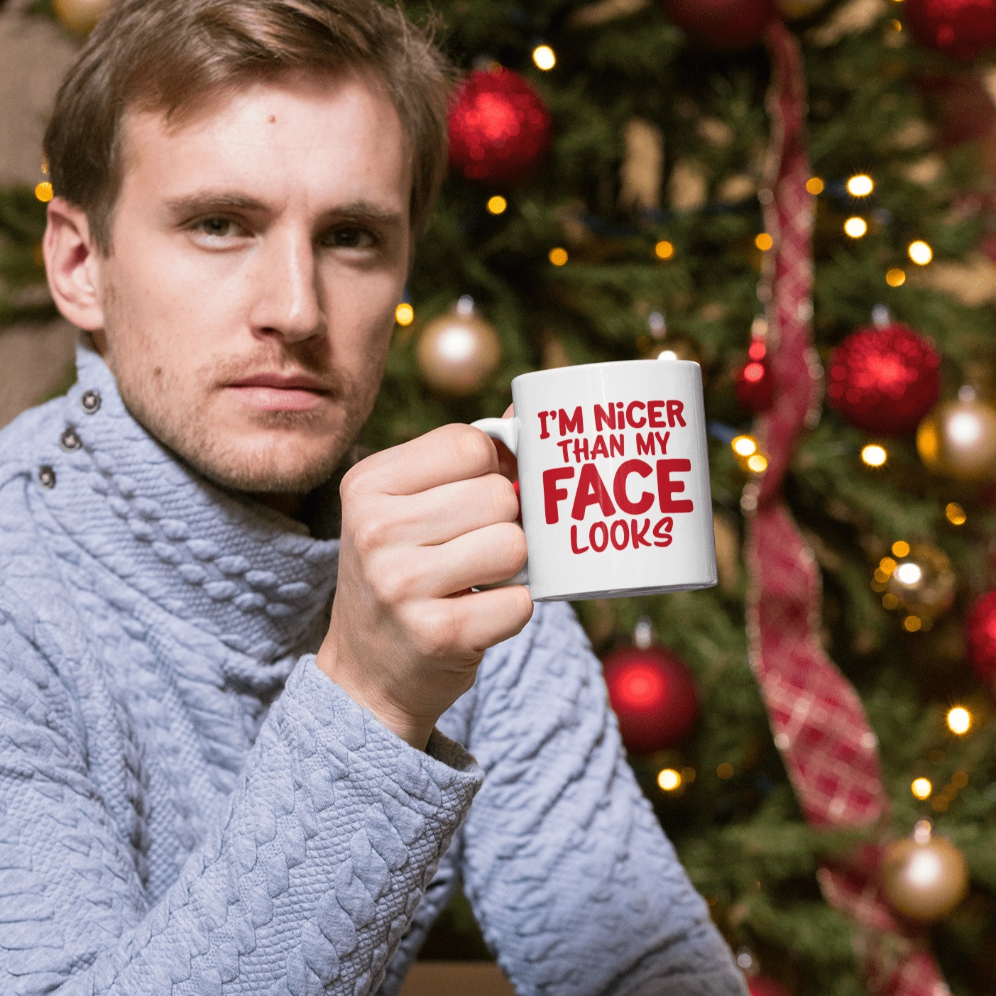 Humorous ceramic mug with "I'm Nicer Than My Face Looks" design, held by man in sweater, Christmas tree background.