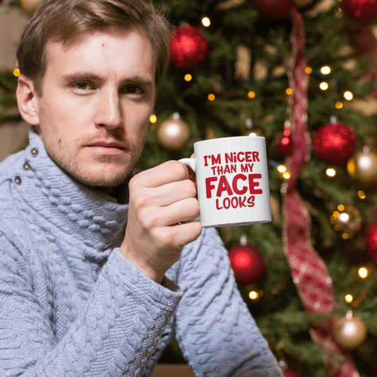 Humorous ceramic mug with "I'm Nicer Than My Face Looks" design, held by man in sweater, Christmas tree background.