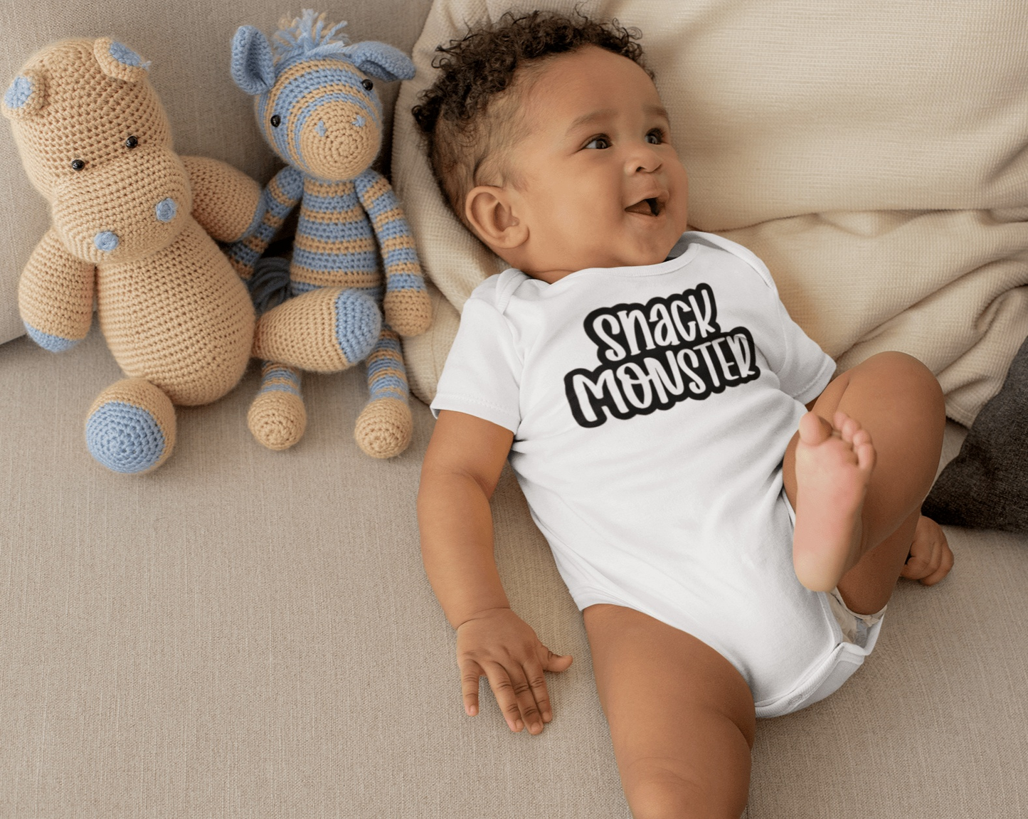 Unisex baby bodysuit "Snack Monster" in white cotton with baby and plush toys on a couch.