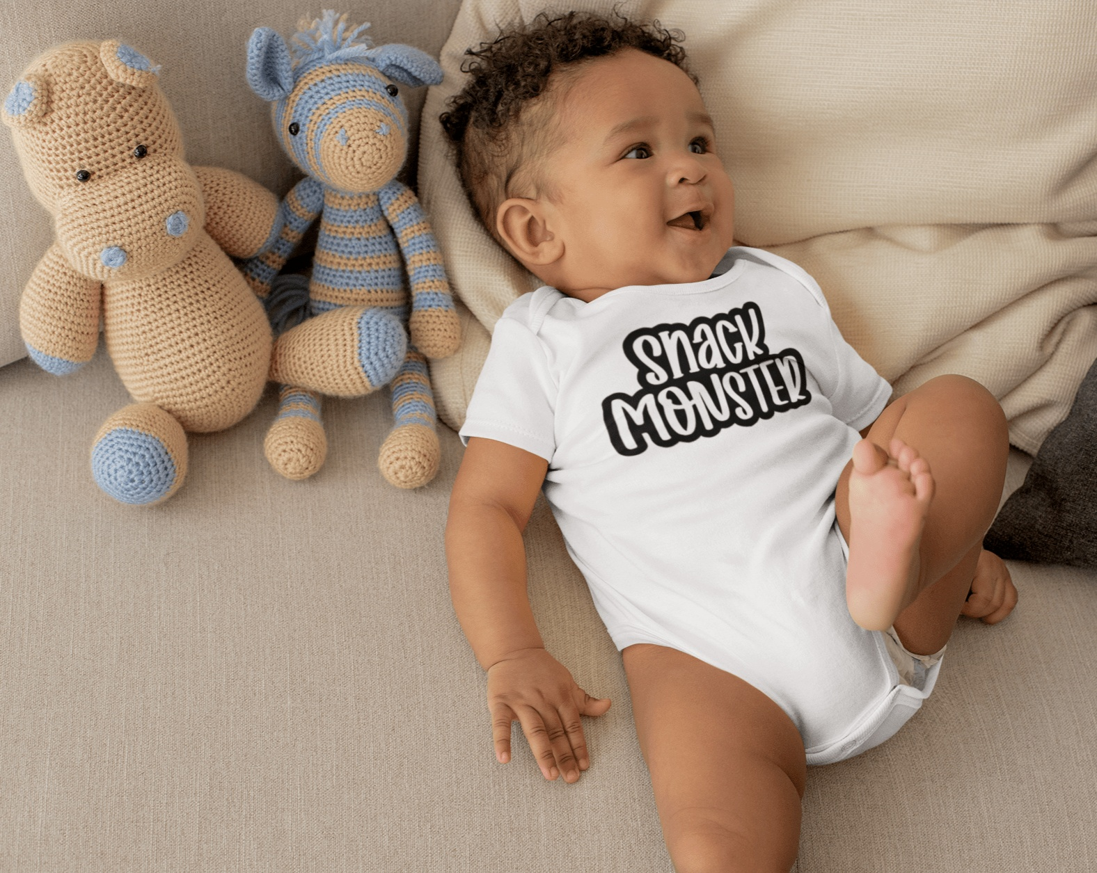 Unisex baby bodysuit "Snack Monster" in white cotton with baby and plush toys on a couch.