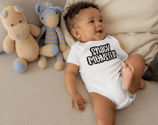 Unisex baby bodysuit "Snack Monster" in white cotton with baby and plush toys on a couch.