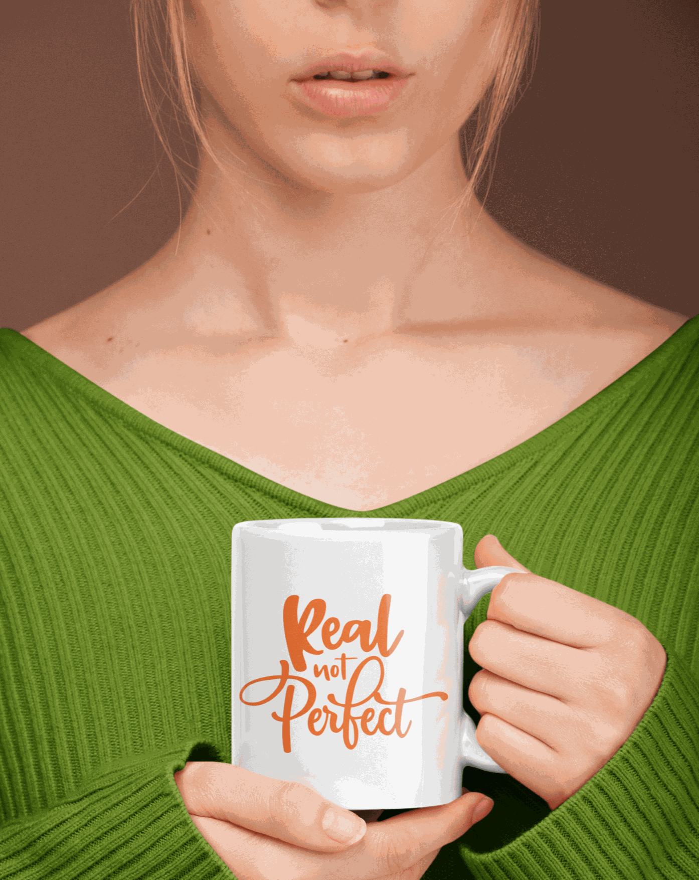Ceramic coffee mug with "Real Not Perfect" text in orange, held by a person in a green sweater.