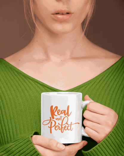 Ceramic coffee mug with "Real Not Perfect" text in orange, held by a person in a green sweater.