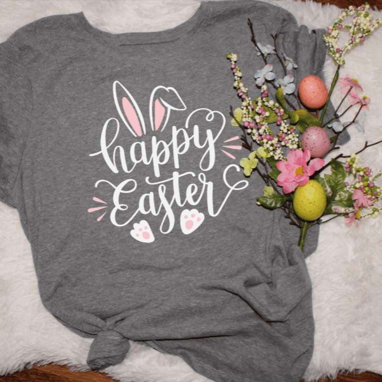 gray women's Happy Easter shirt with bunny ears and paw prints design beside pastel Easter eggs and spring flowers