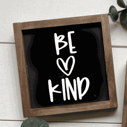 Handmade "Be Kind" sign with black background and white lettering in wooden frame.