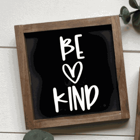 Handmade "Be Kind" sign with black background and white lettering in wooden frame.