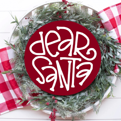 Dear Santa wood sign with white lettering on a red round pine background, surrounded by holiday greenery and checkered fabric.