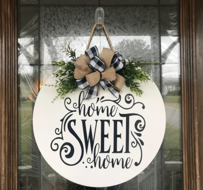 Round wooden "Home Sweet Home" sign with decorative ribbon and greenery hanging on a front door.