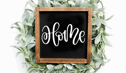 Handcrafted wood sign with "Home" text on black background, framed in wooden border, surrounded by greenery.