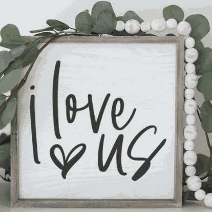 Handcrafted I Love Us wood sign with black lettering on white background, framed in stained pine wood, surrounded by greenery.