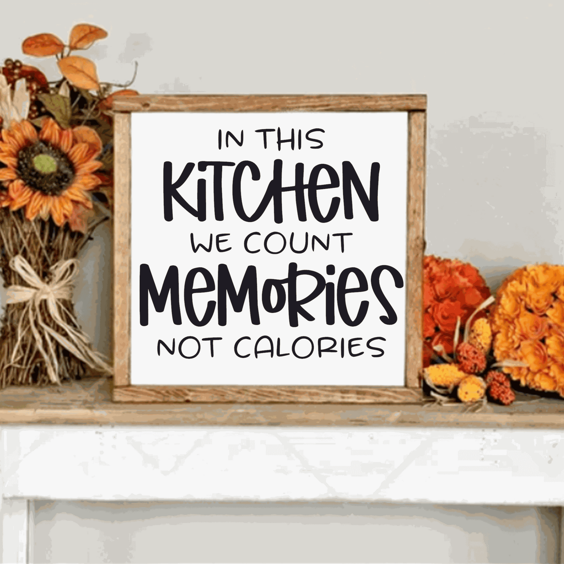 In this Kitchen we Count Memories not Calories wood sign with black lettering on a white background, framed, displayed with floral decor.