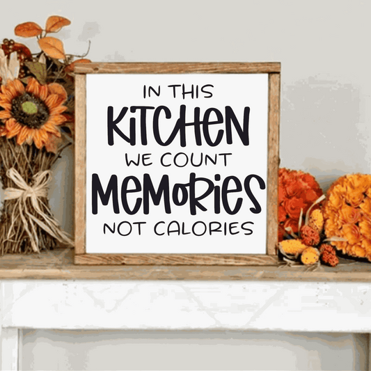 In this Kitchen we Count Memories not Calories wood sign with black lettering on a white background, framed, displayed with floral decor.