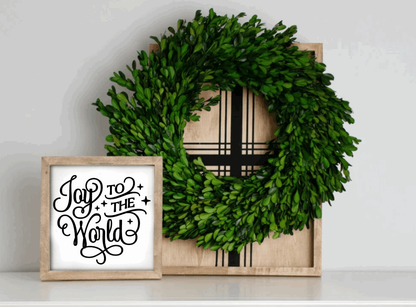 Handmade wood framed sign with "Joy to the World" text in black on an antique white background, sitting by a green wreath.